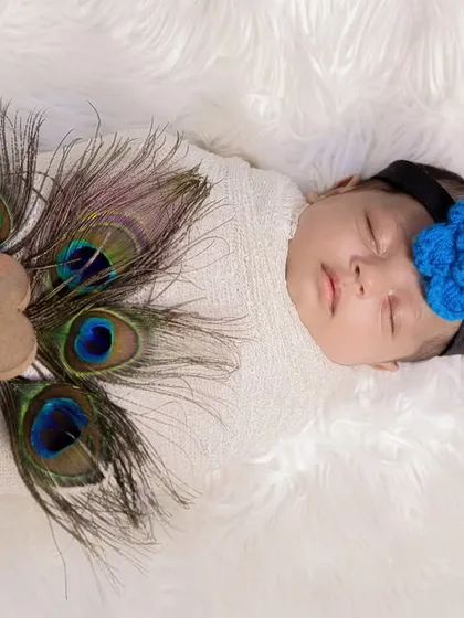 A newborn wrapped in white, adorned with peacock feathers and a small wooden heart. This is a simple yet elegant nod to the Krishna theme.