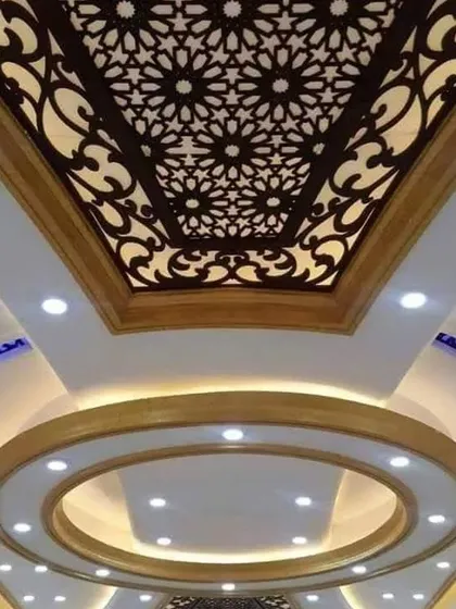 A grand ceiling that combines a circular recessed section with a large, ornate jali panel. The mix of spotlights and cove lighting creates a bright and luxurious space.