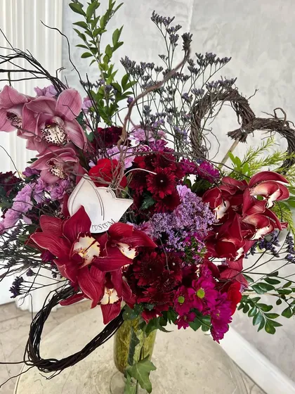 Love first, flowers second, kindness always. This Valentine's arrangement combines deep red Cymbidium orchids with roses and chrysanthemums, finished with wild, woody foliage for a rustic, romantic feel.