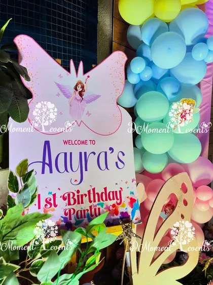 A close-up of a custom welcome sign for Aayra's first birthday party. The sign is shaped like a butterfly and surrounded by lush greenery and colorful balloons.