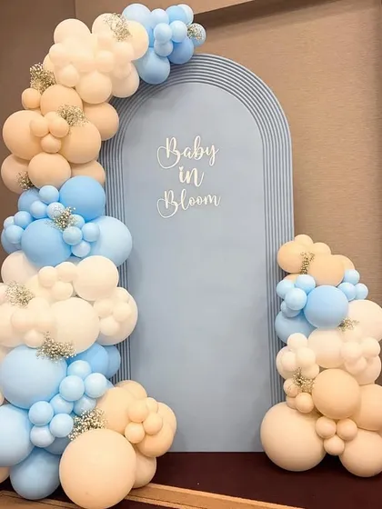 A modern and minimalist 'Baby in Bloom' backdrop for a baby shower. The setup features a stylish blue arched panel framed by an organic balloon garland in pastel blue, white, and beige.