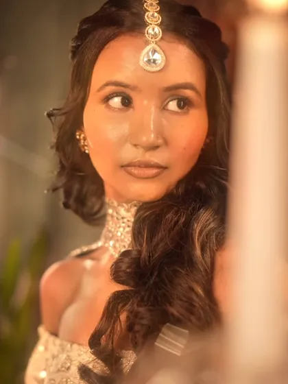 A close-up portrait from the same shoot, with dramatic, warm lighting. The makeup is designed to look flawless and glowing on camera.