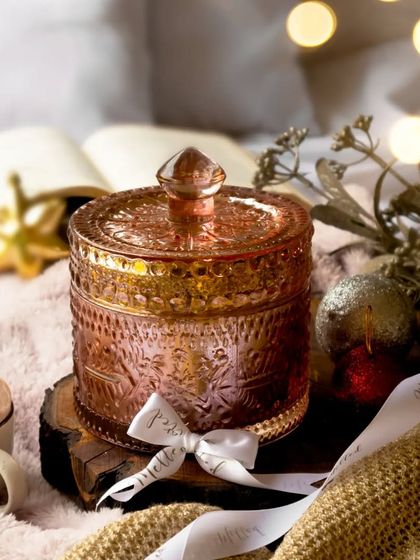 A beautiful pink glass candle jar sits in a cozy winter scene with soft blankets, a book, and festive lights. The warm tones and soft textures create a feeling of holiday comfort and relaxation.