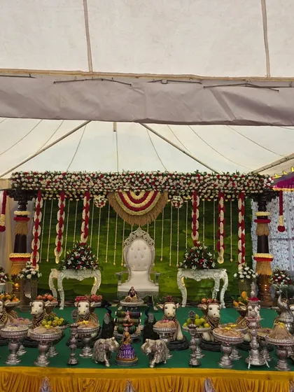 A traditional decor setup inside a large tent, featuring a green backdrop with red and white floral garlands.