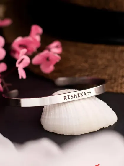 A silver cuff bracelet engraved with 'RISHIKA' and a small symbol, resting on a seashell. This product shot highlights the option to add small icons alongside a name.