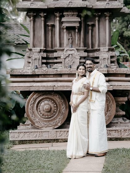 A formal portrait of a couple in traditional Kerala attire, posing in front of a stone temple chariot replica.