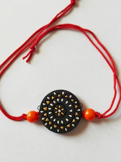 This rakhi features a delicate spiral pattern in white and yellow on a black base. It's a simple yet striking design, attached to a traditional red thread.
