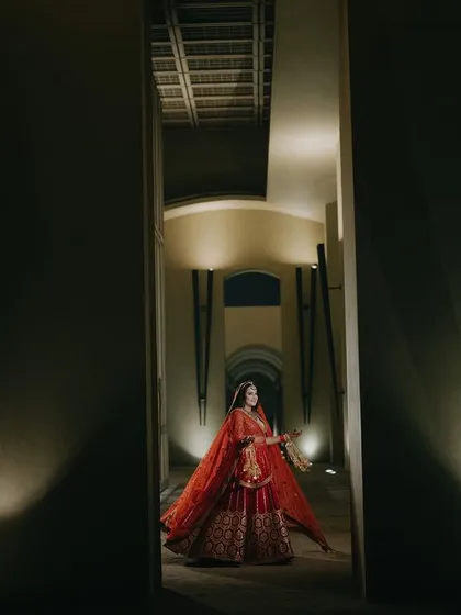 A grand portrait of Tamanna in her red bridal lehenga, framed by a dark corridor. The lighting highlights her, creating a dramatic and powerful image that emphasizes her stunning attire.