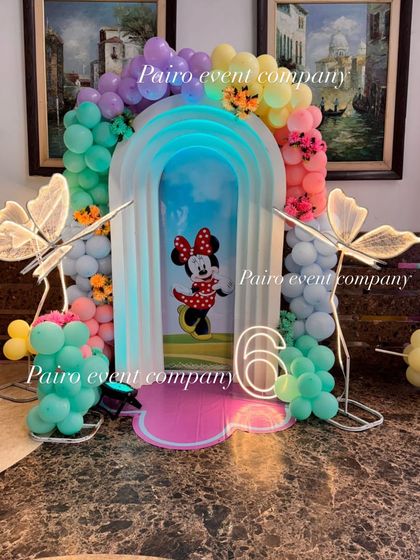 A whimsical Minnie Mouse and butterfly-themed photo booth with a lighted arch, pastel balloons, and a neon number '6'.