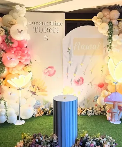 'Our Sunshine Turns 2'. This enchanting butterfly garden backdrop features two balloon arches, a custom name sign, and whimsical light-up mushroom and flower props.