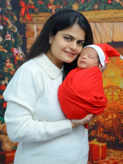 A mother holds her newborn, both dressed for the holidays in front of a cozy fireplace backdrop. The baby wears a tiny Santa hat, making for a perfect first Christmas memory.