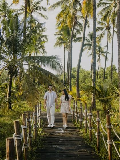 The couple walks hand-in-hand down a rustic wooden path surrounded by palm trees. This shot has a serene, tropical resort feel, perfect for a destination pre-wedding shoot.