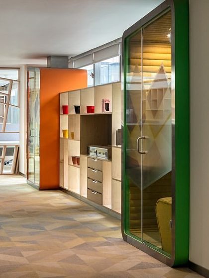 A soundproof telephone booth at the Stainless Steel Office provides a private space for calls. The vibrant green and orange colors distinguish it from the main workspace, adding a playful yet functional element to the office floor.