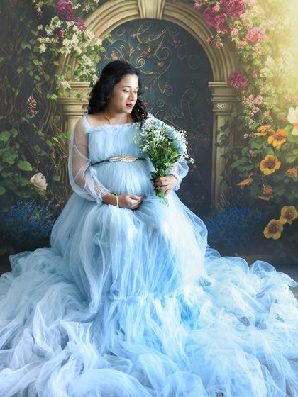 A dreamy, Cinderella-like portrait. The mom-to-be is sitting in a light blue gown, holding a bouquet, against our floral arch backdrop.