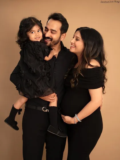 A classic family portrait in the studio. The parents and their daughter, all dressed in black, smile lovingly at each other, creating a timeless and elegant family maternity photo.