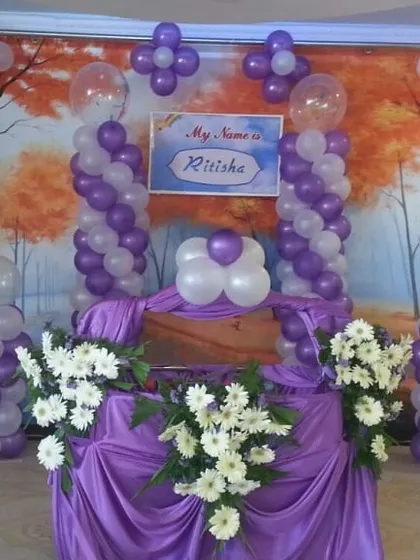 A simple at-home naming ceremony setup with a purple and white color scheme. The cradle is draped in purple fabric, surrounded by balloon pillars and fresh white flowers.