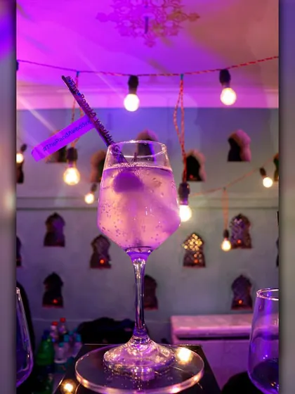 A sparkling, lavender-hued cocktail served in an elegant wine glass. The subtle glow from the background lights enhances its magical appearance.