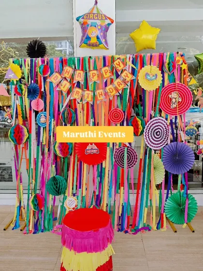 A colorful circus or carnival-themed party. The backdrop is made of vibrant streamers and paper fans, with a fringed plinth adding to the festive look.