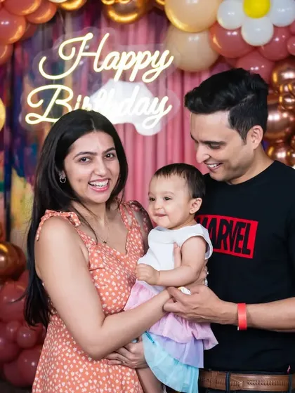 A perfect family portrait capturing the parents and their one-year-old daughter smiling in front of a neon 'Happy Birthday' sign.
