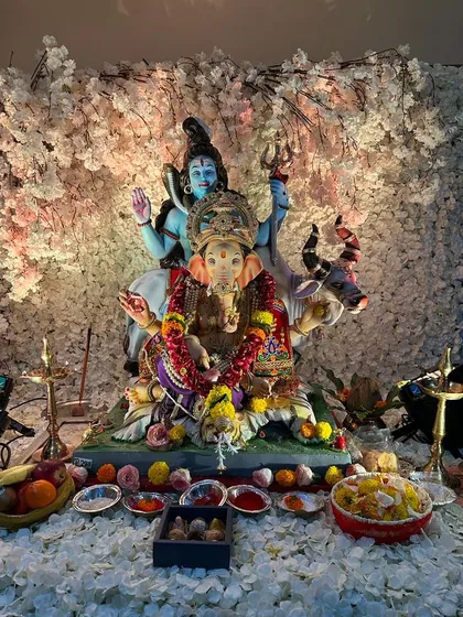 An elegant and serene home Ganpati setup featuring a beautiful idol of Ganesha with Shiva and Parvati. The backdrop is a dense wall of delicate white blossoms, creating a heavenly and peaceful ambiance for prayer and offerings.