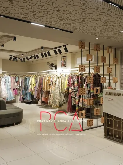 An overall view of the Jubinav Chadha fashion studio, showing the reception desk, customer seating area, and extensive clothing displays. The design balances open space for browsing with intricate details inspired by Indian temple architecture.