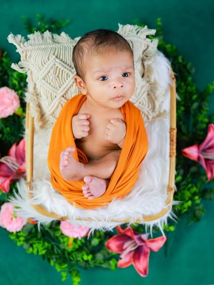 This little one is awake and looking around with such curiosity. The vibrant orange wrap contrasts beautifully with the green floral wreath, creating a striking portrait.