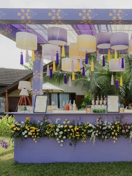 A chic outdoor cocktail bar designed for a pastel-themed Haldi. The lavender-colored bar is adorned with fresh flowers and features an overhead canopy with hanging lampshades and colorful tassels.