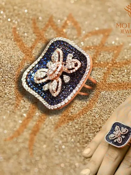 A striking butterfly-inspired ring with blue and white diamonds, set in rose gold, resting on a bed of sand.