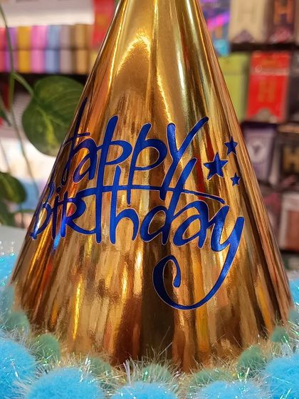 A shiny gold birthday hat with blue 'Happy Birthday' text and a fluffy blue pom-pom border. This is a fun and festive accessory for the birthday person to wear on their special day.