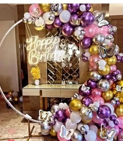A birthday party with a circular frame and a balloon garland in purple, pink, gold, and silver. The setup includes a "Happy Birthday" neon sign and butterfly accents.