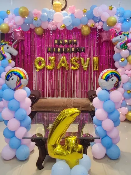 A unicorn-themed fourth birthday party using a pink, blue, and gold color palette. The decoration includes a full balloon arch over a couch, a shimmer backdrop, and several unicorn and rainbow foil balloons.
