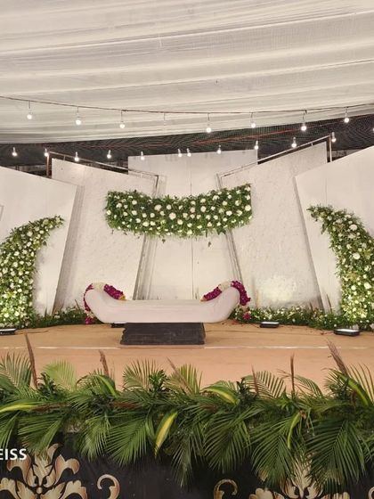 A sophisticated all-white reception stage. The design uses angled panels and cascading white floral arrangements to create a modern and architectural look.