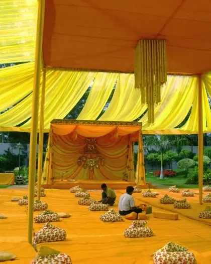 A large-scale Satsang or Haldi setup under a bright yellow tent. The space is arranged with low floor seating using printed cushions, all oriented towards a decorated stage, perfect for a spiritual or traditional gathering.