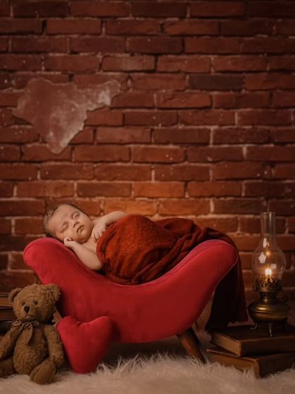 A newborn sleeps peacefully on a miniature red armchair against a rustic brick wall, with a teddy bear and an old lantern as props.