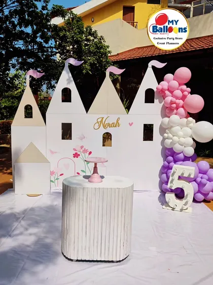 A life-sized castle backdrop for a fifth birthday. This simple yet elegant setup features a large white castle cutout, a single dessert plinth, and a purple and pink balloon garland.