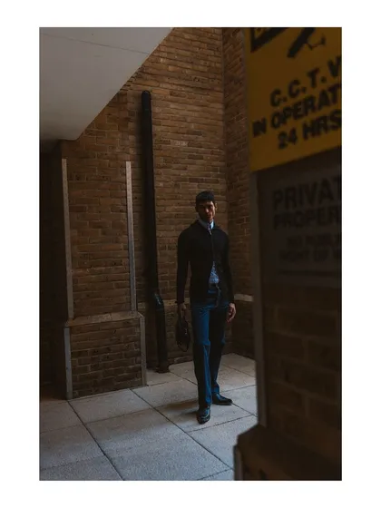 A full-length shot in an urban alleyway. The composition uses the surrounding walls and signs to frame the model, creating a gritty and atmospheric menswear editorial image.