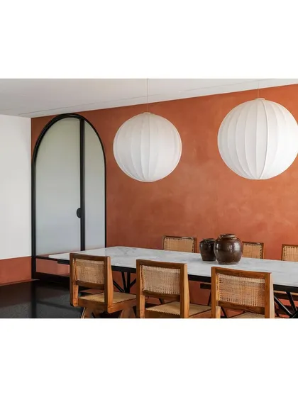 The dining space in the Ved House features a textured terracotta wall, a cool marble tabletop, and warm cane chairs. This interplay of authentic materials and colors creates a rich, inviting atmosphere for meals.