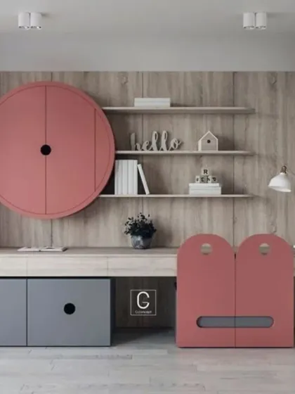 A playful and modern study table design for a child's room. The unit features unique circular and arched cabinet doors in pink and grey, open shelving, and a long desk surface.