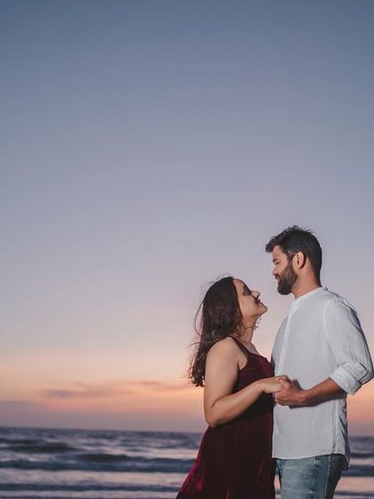 An intimate moment between a couple, holding hands and looking at each other against the backdrop of a soft sunset over the sea.