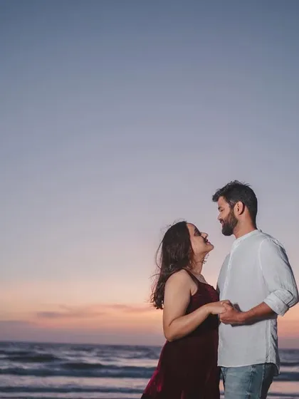 An intimate moment between a couple, holding hands and looking at each other against the backdrop of a soft sunset over the sea.