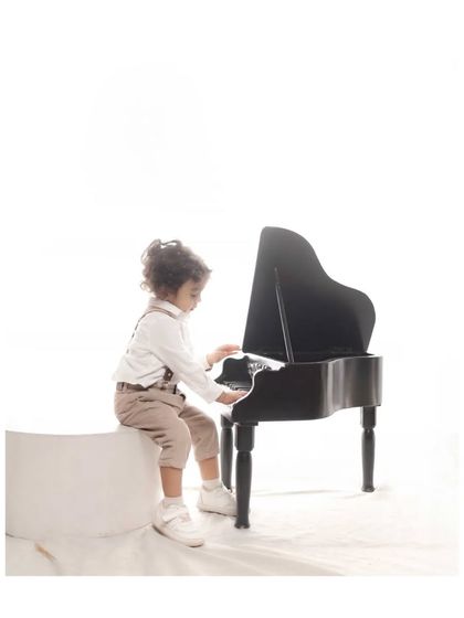 A little boy dressed in smart trousers and suspenders sits at a miniature grand piano. This classic and charming studio portrait captures the timeless innocence of childhood with a touch of old-world style.