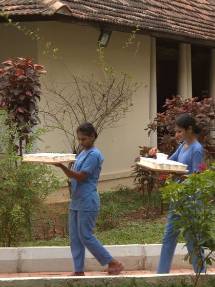 Two of our therapists walk through the garden carrying trays of herbal preparations for guests. Their work is central to the authentic Ayurvedic experience we provide.
