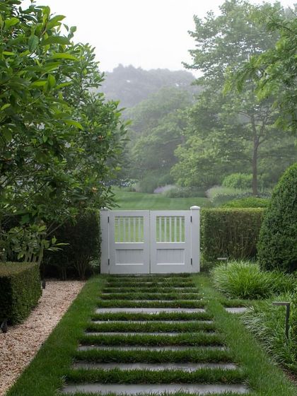 A formal pathway made of stone slabs set into a lawn, leading to a classic white garden gate. The path is bordered by neatly clipped hedges and shrubs, creating a symmetrical and orderly look.