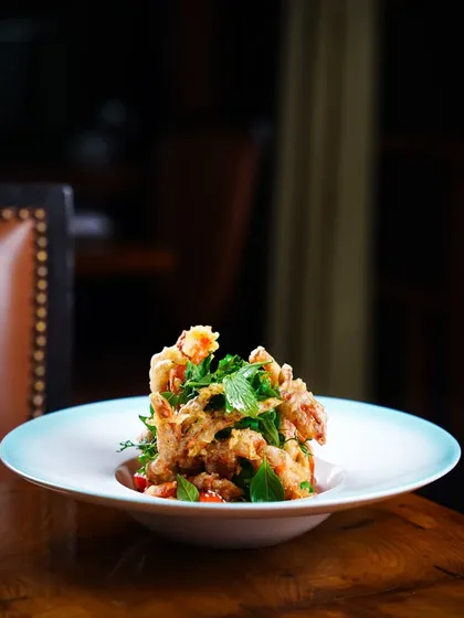 A plate of crispy soft-shell crab, elegantly presented at The Table, Colaba. The dark, moody restaurant interior in the background adds to the sophisticated atmosphere.