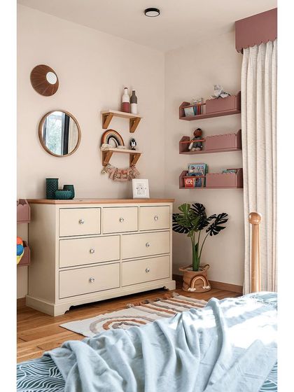 Opposite the bed in this kids' room is a chest of drawers for extra storage. The walls are adorned with open shelves and book ledges featuring scallop details and pastel hues.