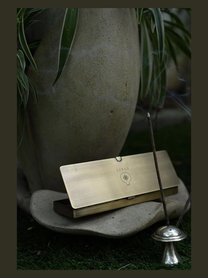 The gentle curl of smoke from an incense stick creates a serene mood. This shot for Byaas Home focuses on their elegant brass incense case, styled in a tranquil green setting to evoke a sense of calm.
