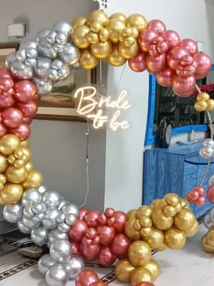 A trendy and modern bachelorette party photo booth featuring a large circular balloon arch. The arch is made of metallic rose gold, gold, and silver balloons, with a "Bride to be" neon sign glowing in the center, perfect for a stylish home party.