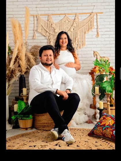 A couple's portrait in a bohemian-themed studio, with the mom-to-be in our white one-shoulder ruffled gown.
