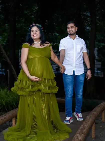 An elegant maternity portrait of a couple taking a stroll across a small bridge in a park. The flowing olive green gown and the soft, natural lighting create a serene and beautiful scene.