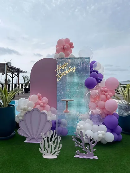 A beautiful mermaid theme birthday decoration set against a poolside lawn. I used a shimmer wall backdrop, a pastel balloon garland in pink, purple, and white, and custom props like a large seashell and coral cutouts.